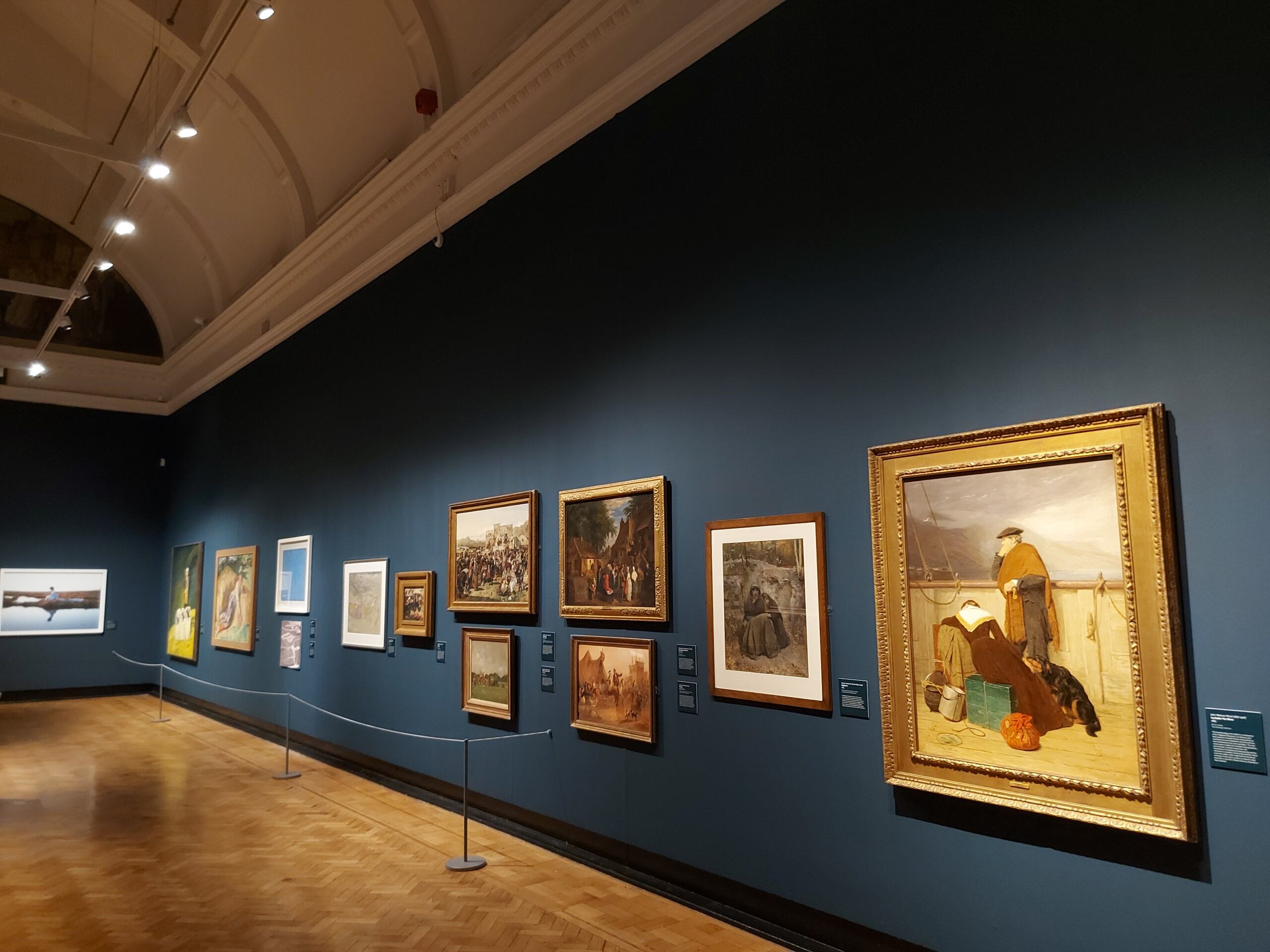 A photograph of an exhibition hall, featuring a line of paintings set against a navy-blue wall. The closest painting depicts a man and a woman gazing pensively from the deck of a ship.