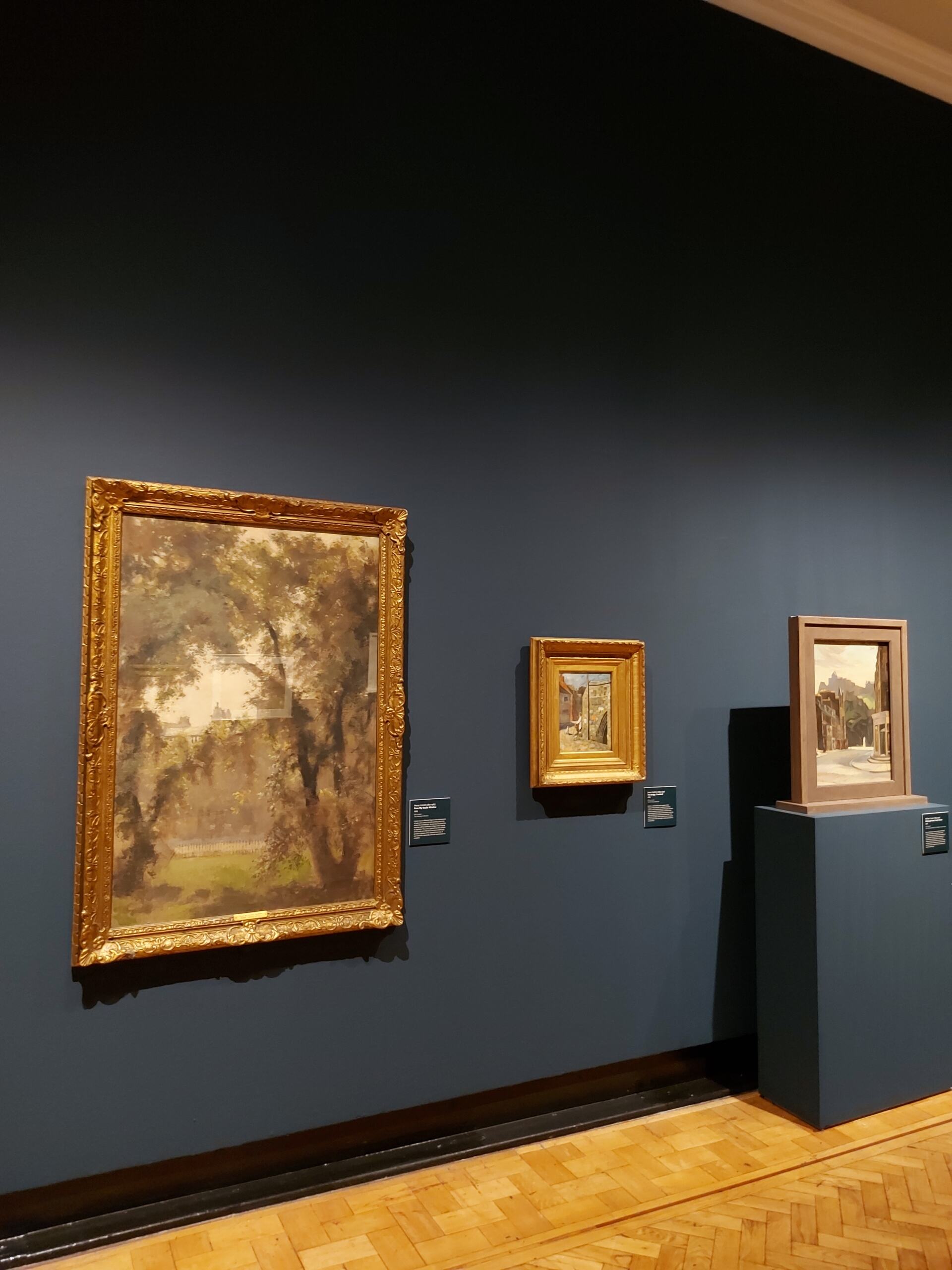 A photograph of an exhibition hall, featuring a line of paintings set against a navy-blue wall. The closest painting depicts a tree arching over a green space with buildings present in the background.