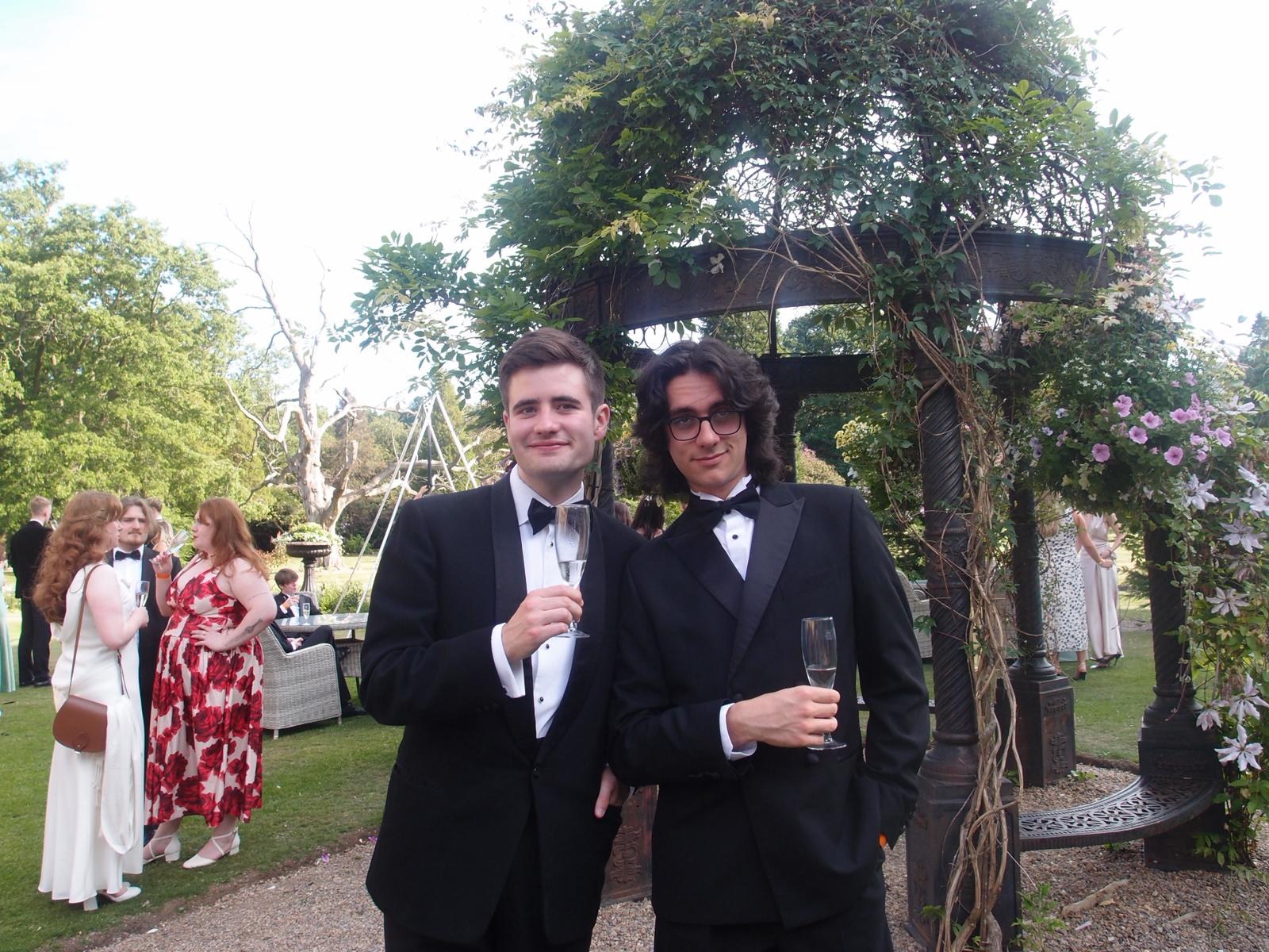 Two rather charming men in black tie pose for a photo. In the background is the gardens of a stately home in the sunshine.