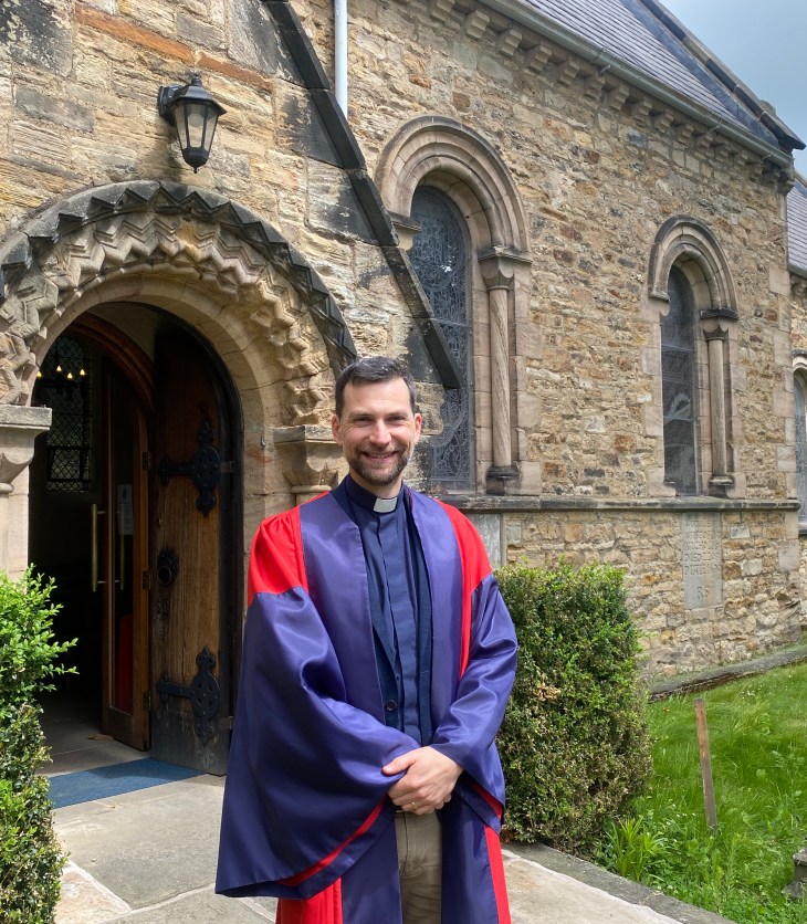 A man wearing a clerical collar and Oxford PhD gown stands outside of a Chapel