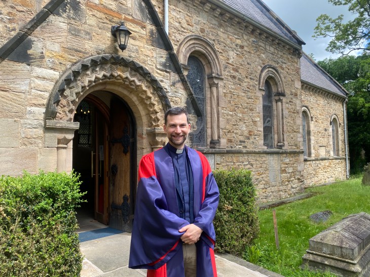 A man wearing a clerical collar and Oxford PhD gown stands outside of a Chapel